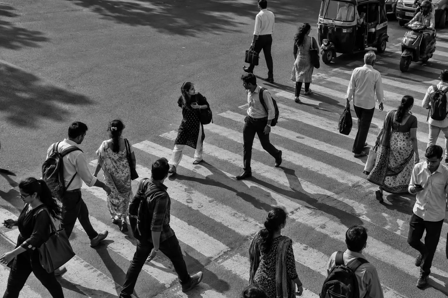 Fictional black-and-white city crossing, with people moving in different directions through hard afternoon shadows.